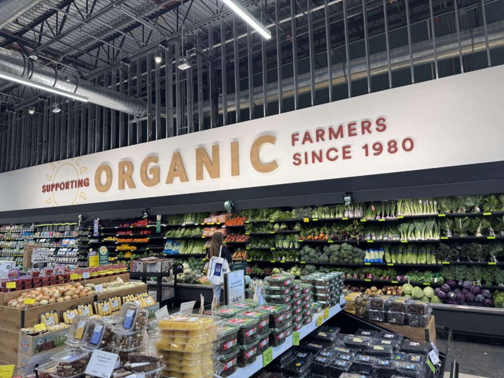 Interior of a grocery store produce section with shelves of fruits and vegetables and an 'ORGANIC' sign in the background