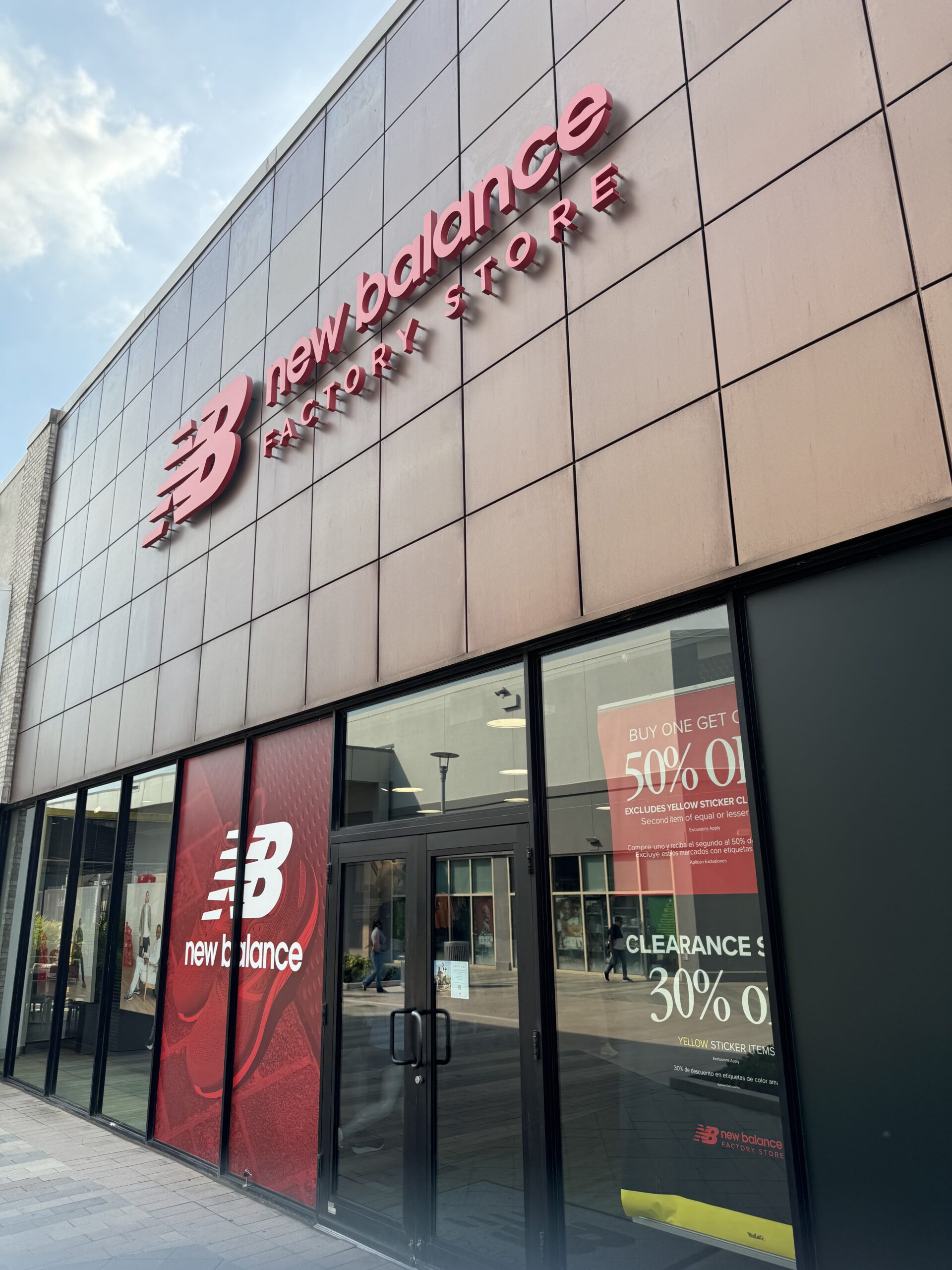 Exterior view of a New Balance Factory Store with a pink-tiled wall and a large red 'NB' logo and 'new balance FACTORY STORE' lettering above glass storefronts with sale posters nearby.