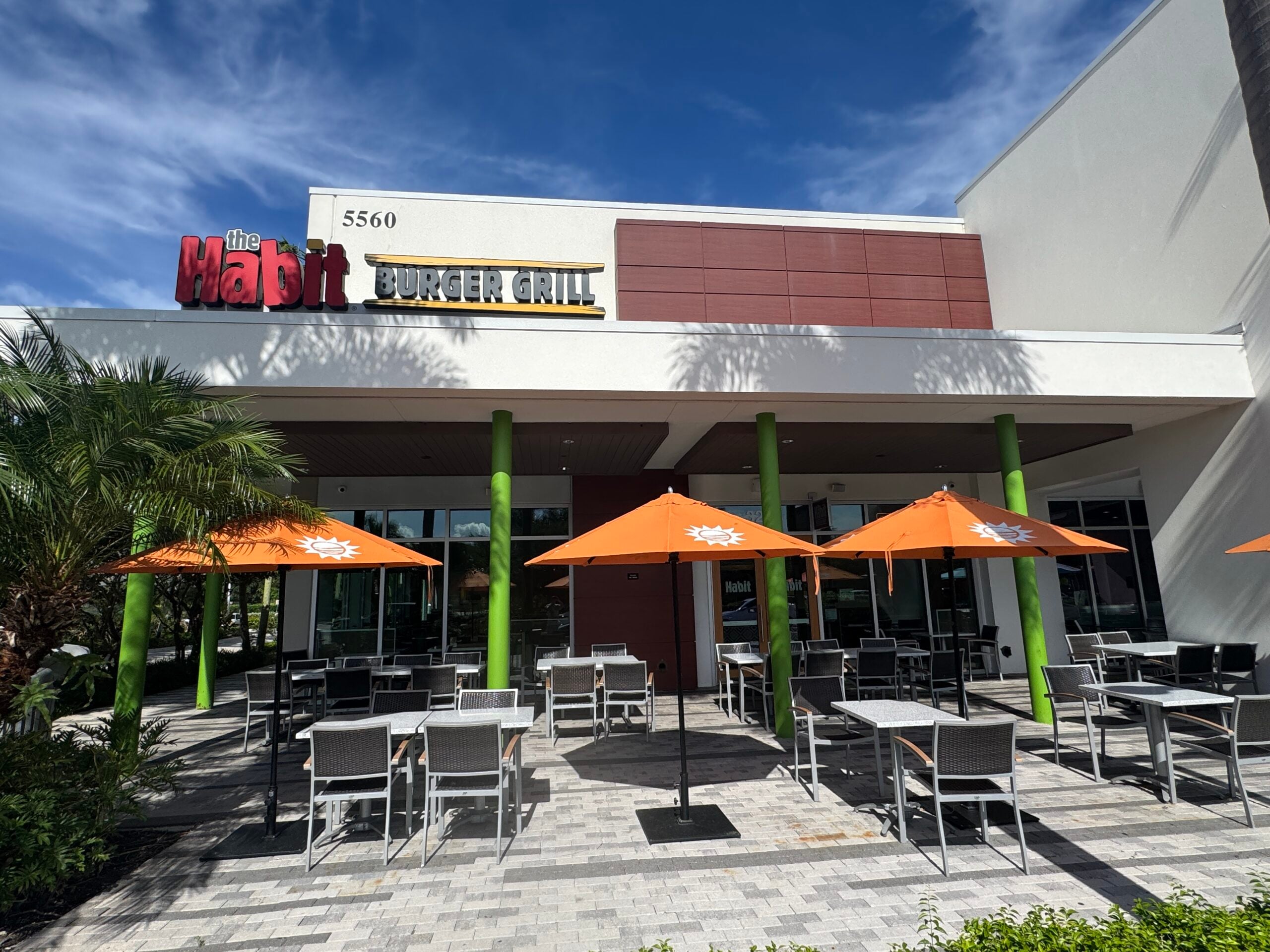 Outdoor dining patio at Habit Burger Grill with orange umbrellas and numerous tables and chairs against a modern white building and blue sky.