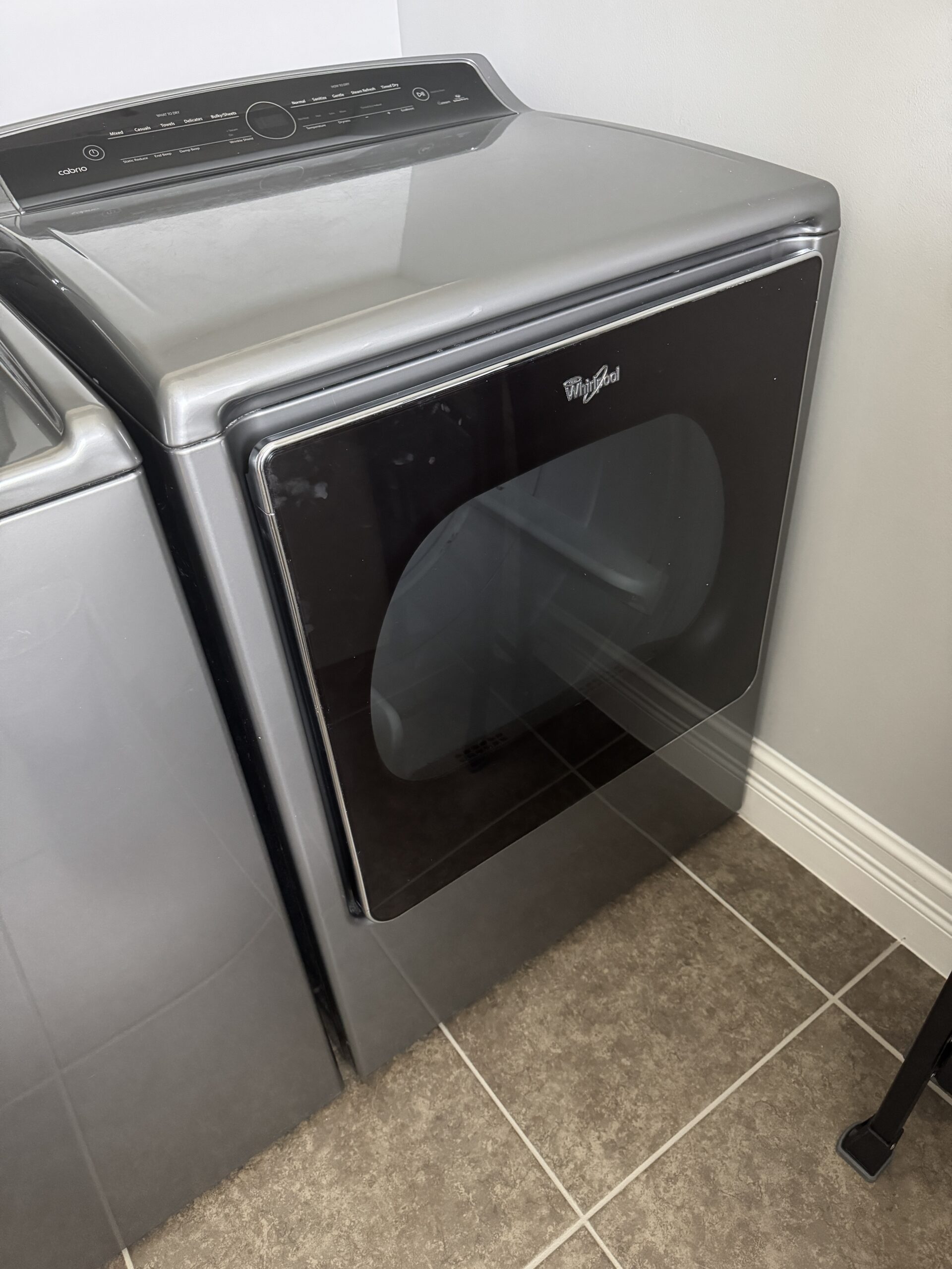 Front-loading Whirlpool washer with a curved glass door and silver control panel, in a laundry room with tiled floor.