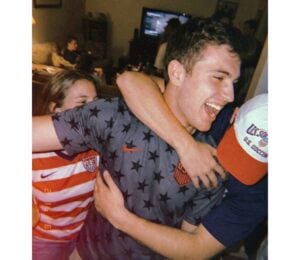 Man in a star-patterned shirt is hugged by friends at a lively indoor gathering, all smiling and cheering.