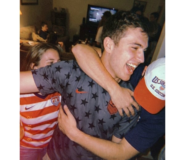 Man in a star-patterned shirt is hugged by friends at a lively indoor gathering, all smiling and cheering.
