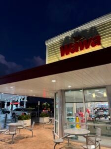 Night view of a Wawa storefront with red logo, illuminated facade, and a covered outdoor seating area with metal tables and chairs under a white canopy