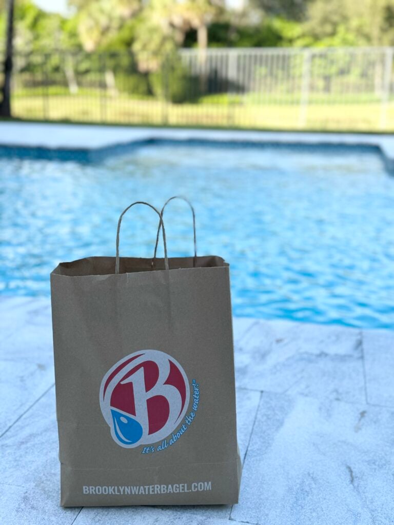 Brown paper bag with Brooklyn Water Bagel logo in foreground, pool and yard in background, outdoor sunny scene, bag sits on a light stone surface.