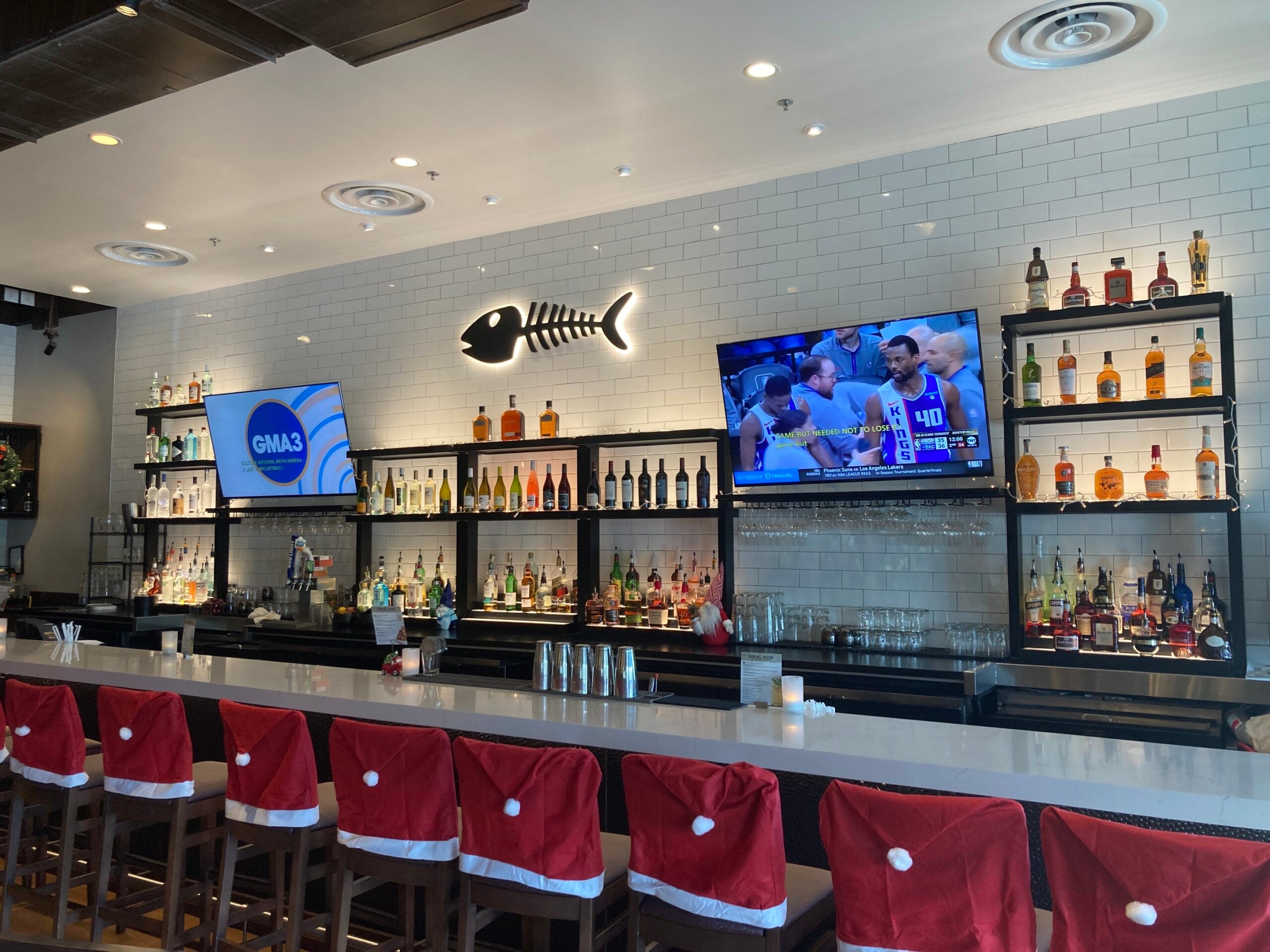 Bar area decorated for Christmas with red Santa-hat stools along a white counter and liquor shelves in the background, plus a fishbone sign on the tiled wall.