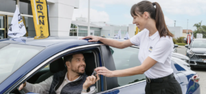 Saleswoman hands car keys to smiling man in a blue sedan at a dealership lot