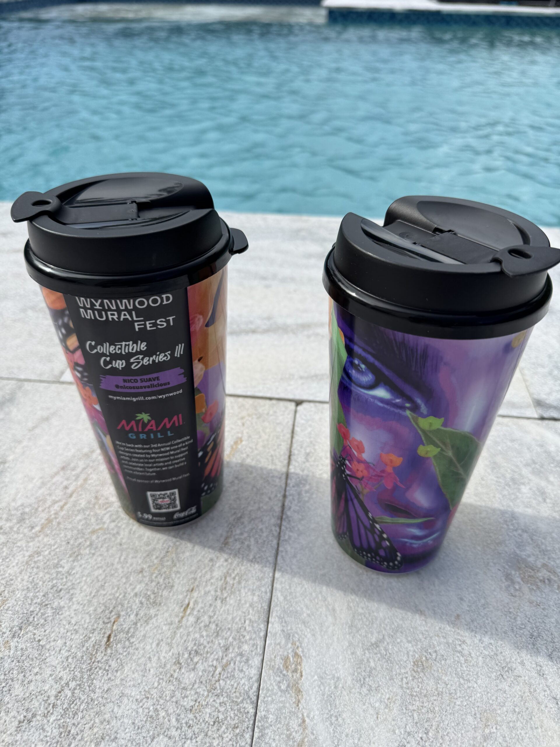 Two colorful travel cups with black lids sit on a poolside surface next to a bright blue pool water edge.