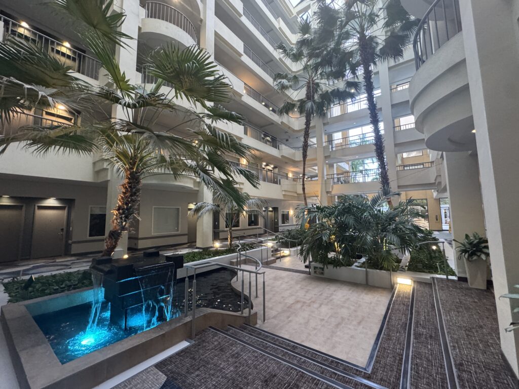 Indoor atrium with palm trees, multiple walkways, and a blue-lit shallow water feature.
