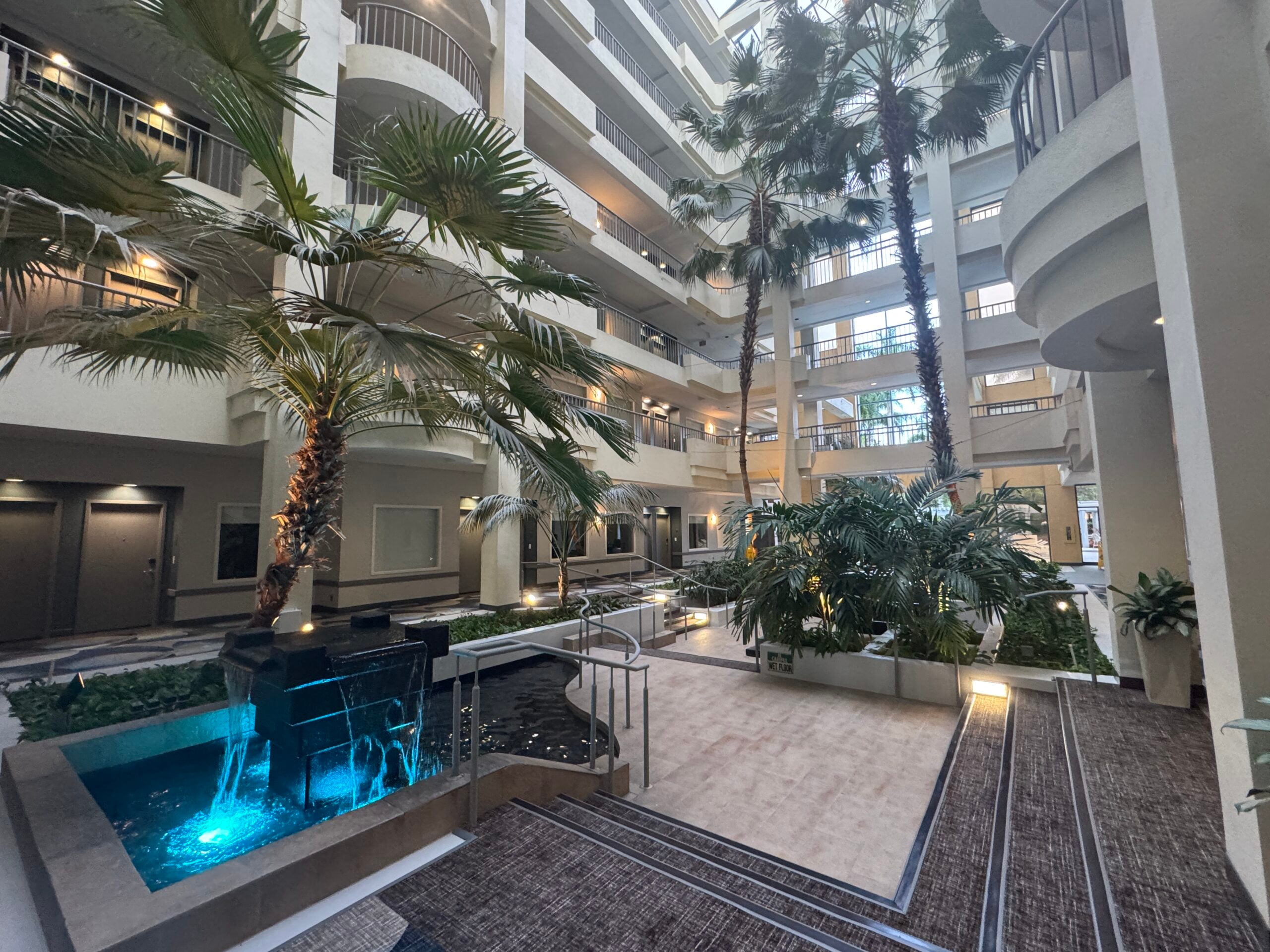 Indoor atrium with palm trees, multiple walkways, and a blue-lit shallow water feature.